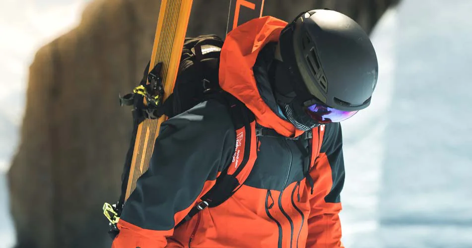 model sa ski oprekom ruksakom i skijama na ruksaku sa kacigom i brilama ski pogledom prema dolje i bijeloj pozadini